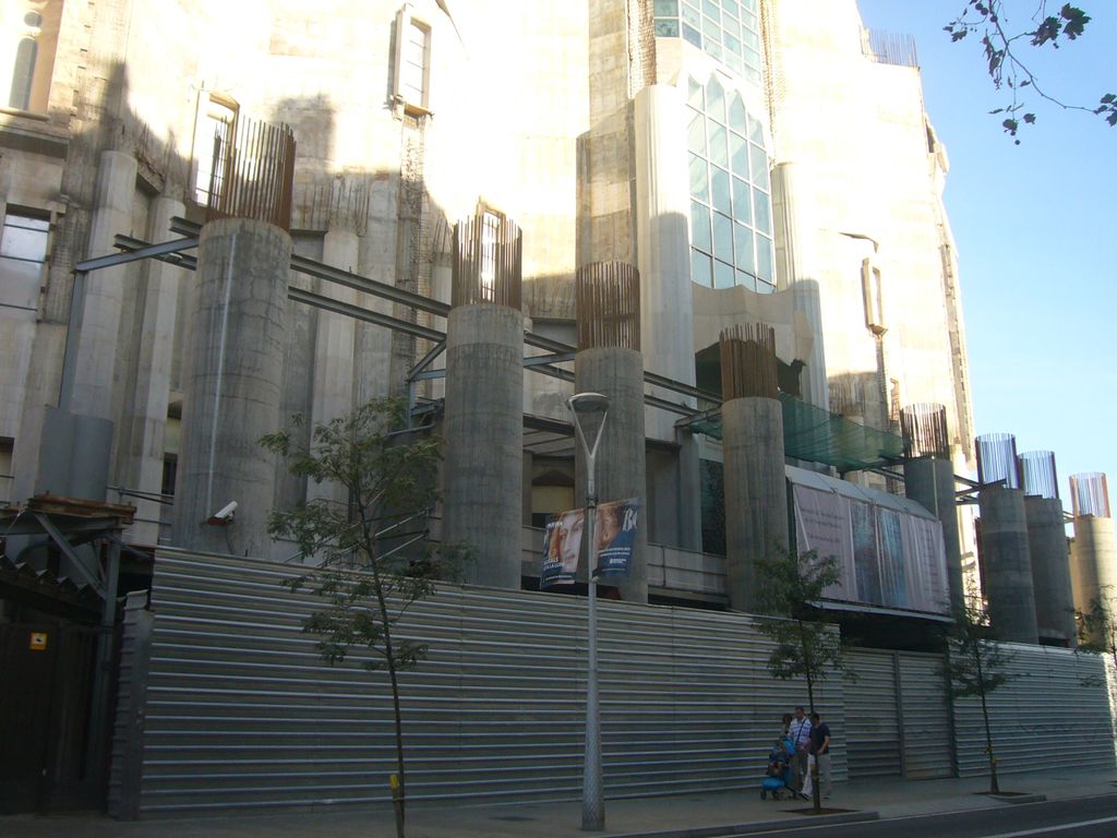 An unfinished church frontage has concrete columns with exposed reinforcing bars, temporary steel framing, partial glazing and a corrugated street fence.