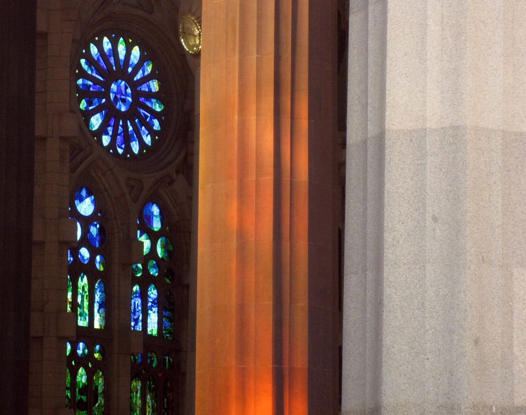 Blue and green stained-glass windows glow beside a massive warm-orange branching column and a pale stone support in a church interior.