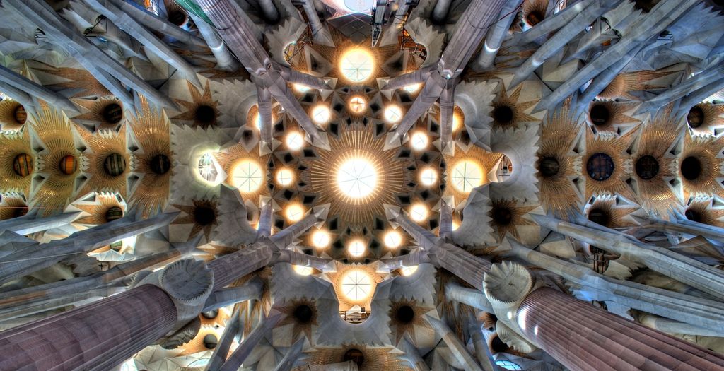 A symmetrical upward view shows a bright circular central opening ringed by many smaller light wells, folded vault surfaces and branching stone supports.