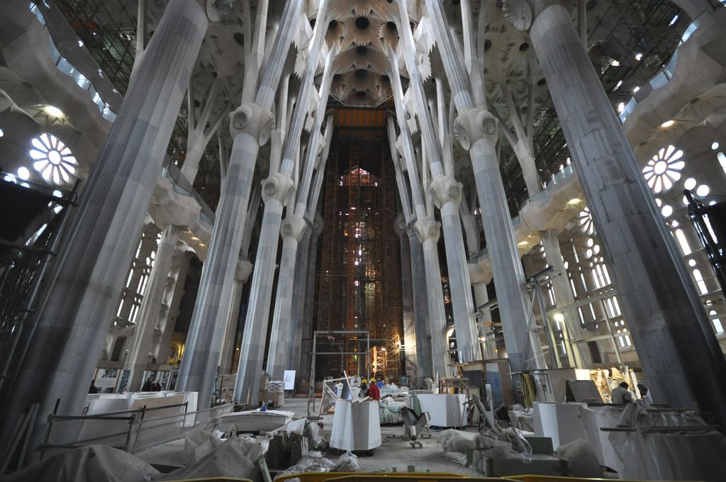 Branching stone columns rise through a broad unfinished church interior filled with blocks, worktables, barriers, scaffolding and workers.
