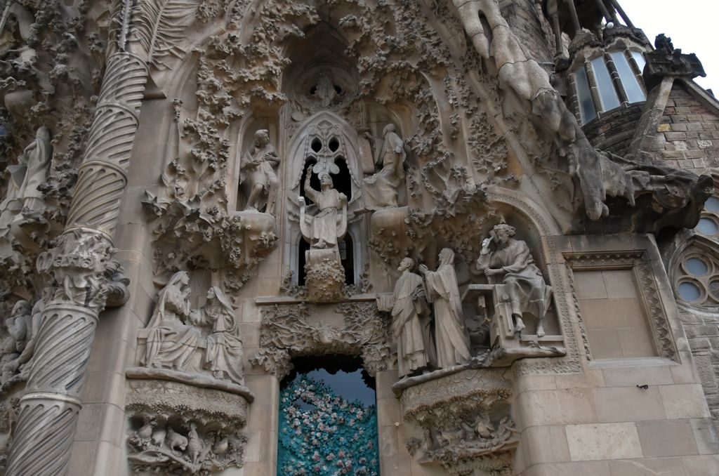 A dense pale-stone façade detail combines many carved figures, leaves, birds, rocky textures, a spiral column and a doorway covered with blue-green flowers.