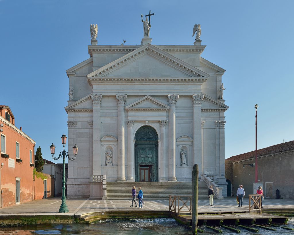 A bright white church façade combines several overlapping triangular pediments, Corinthian columns, statues and a tall stair beside a narrow canal edge.