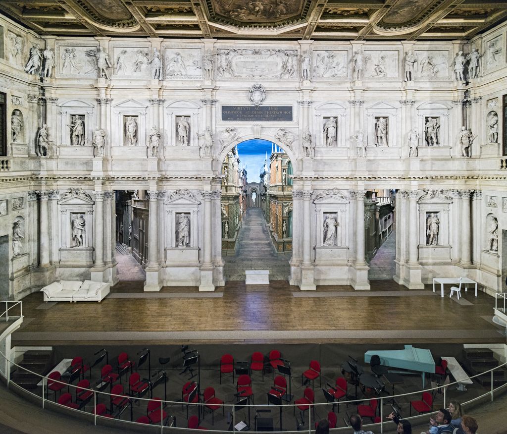 An elaborate columned stage wall with statues opens onto a long painted perspective street, while red chairs and modern equipment fill the orchestra area below.