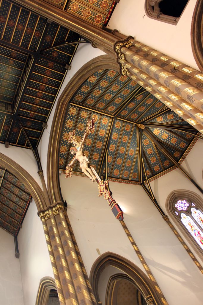 An upward church view shows a dark blue and gold painted timber ceiling, patterned columns and arches, a suspended crucifix and a bright stained-glass window.