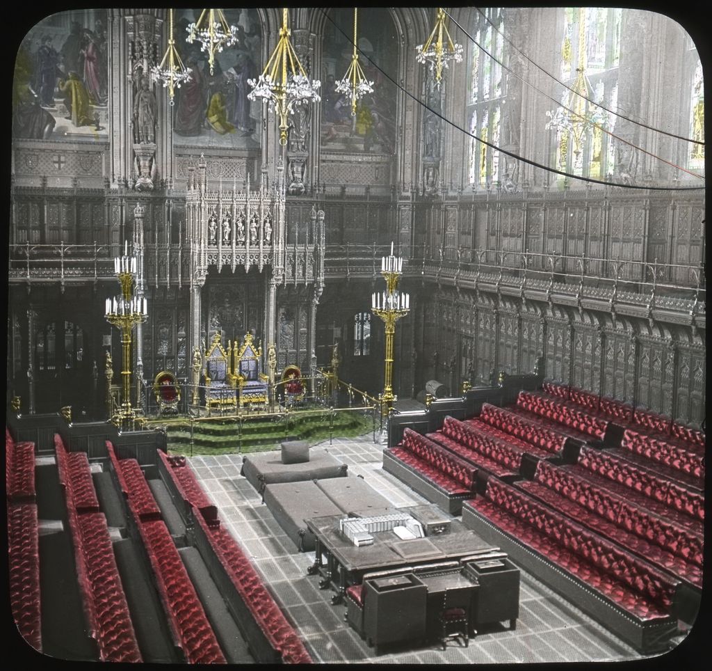 A 1913 colour lantern slide looks down into the House of Lords chamber, with a gilded throne, red facing benches, tall windows, chandeliers, wall paintings and elaborate dark Gothic woodwork.