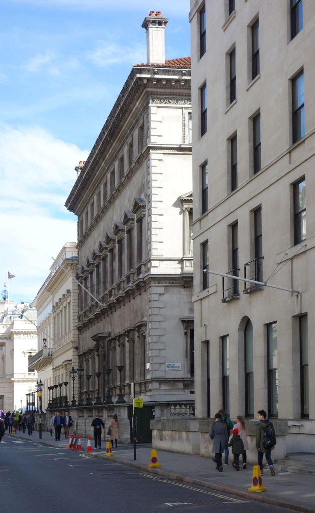 A long, restrained stone club façade with a deep cornice, rusticated corner and regular rows of framed windows recedes along a narrow London street beside a modern building.