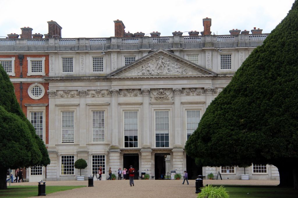 A pale pilastered palace front with a sculpted pediment and tall windows is flanked by red-brick ranges and clipped yew trees in a formal garden.