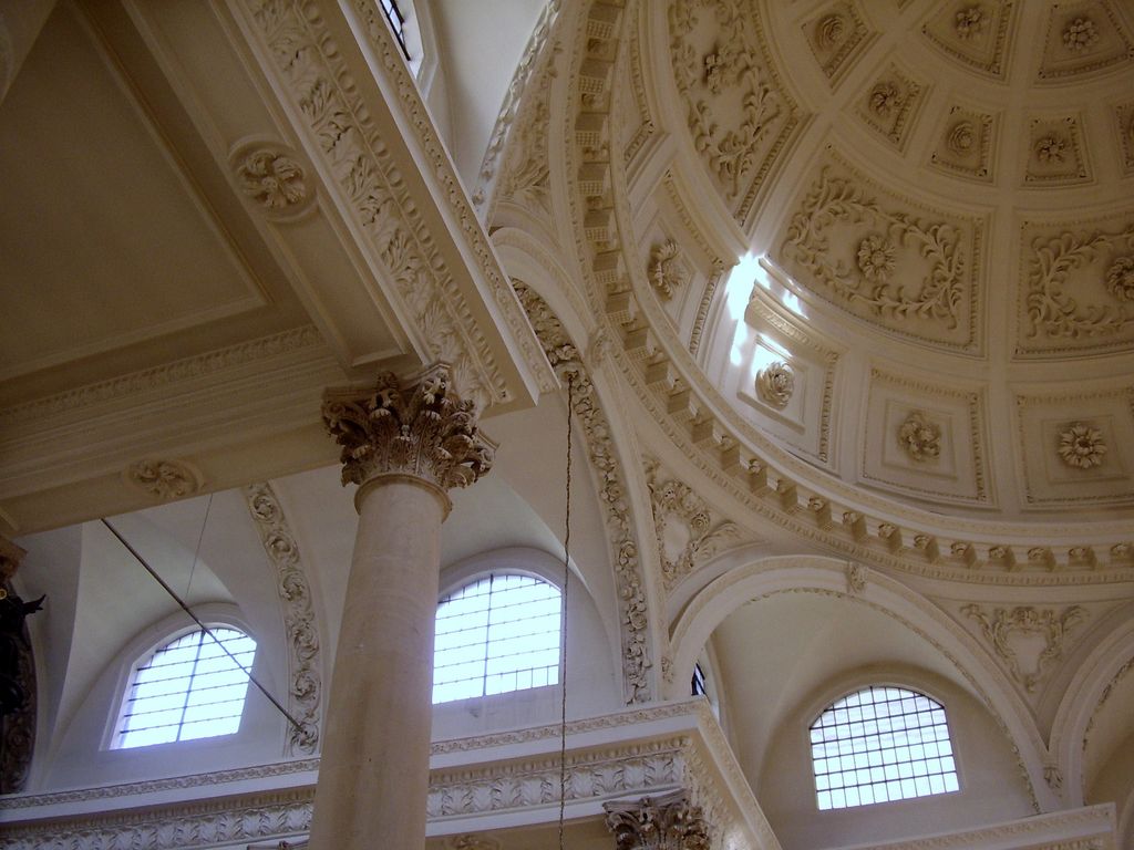 Pale Corinthian columns and richly moulded arches rise towards a coffered dome and high semicircular windows in a bright church interior.