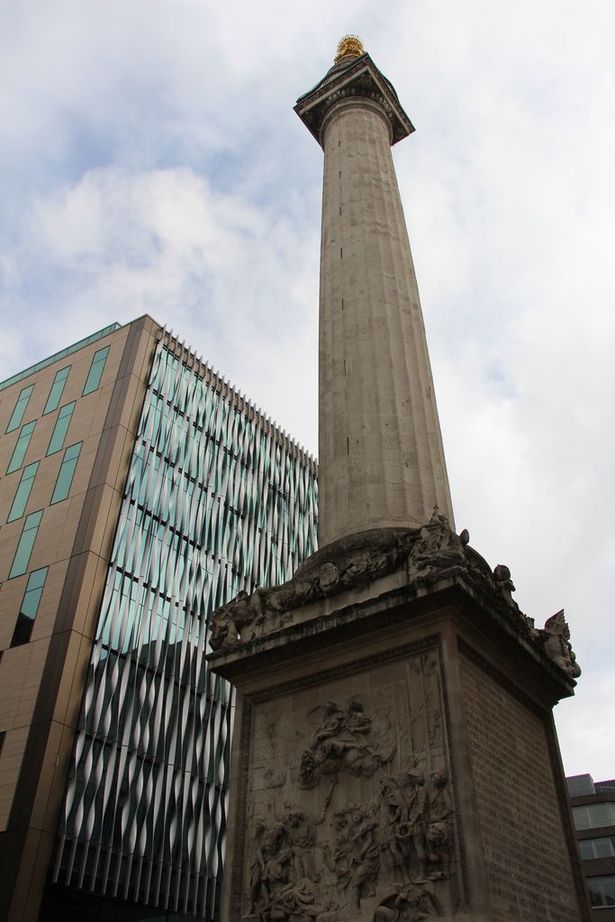A very tall fluted stone column rises from a relief-covered square base to a viewing platform and small gilded flaming urn beside a modern glazed building.