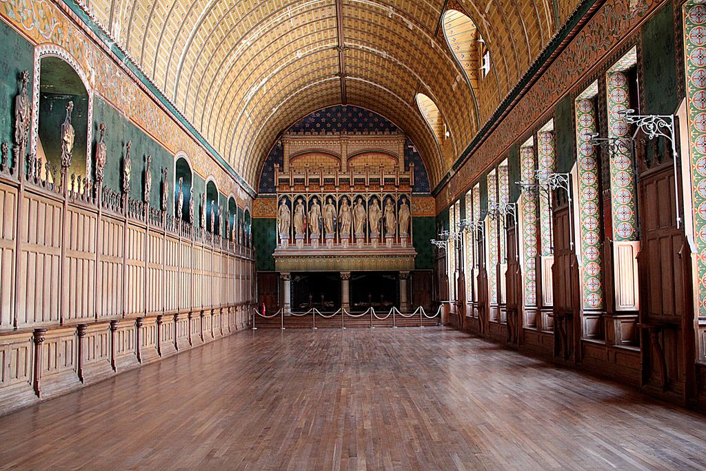 A long richly coloured hall has a high curved timber ceiling, green and gold painted walls, repeated carved figures, wood panelling, patterned windows and a theatrical display at the far end.