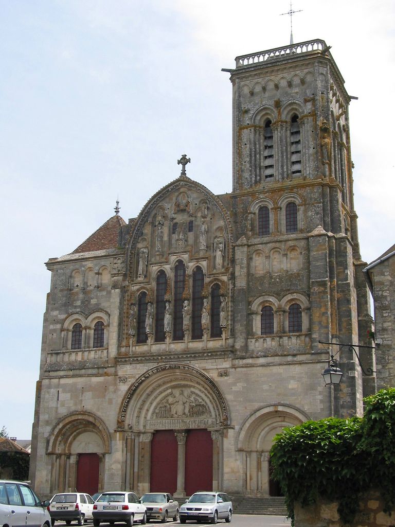 A tall weathered Romanesque church façade has three arched portals, a richly sculpted central tympanum, grouped upper windows and unequal side towers, with cars parked below.
