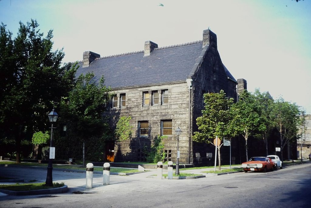 A severe gray granite house with a steep dark roof and few street-facing windows occupies a corner behind trees, lamps and parked 1980s cars.