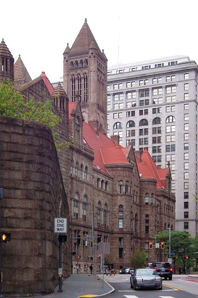 A large rusticated stone courthouse with red roofs, clustered round turrets, rows of arched windows and a tall square tower stands among modern downtown buildings and traffic.