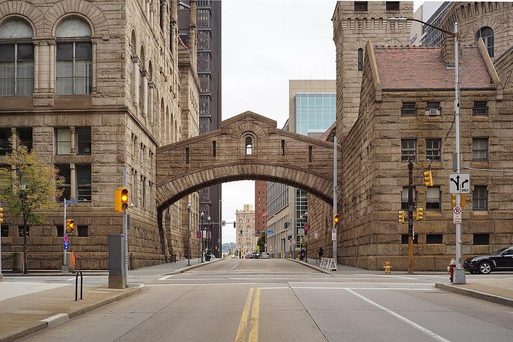 A heavy enclosed stone bridge with a broad round arch crosses an empty city street between two rusticated courthouse and jail buildings amid traffic lights and signs.