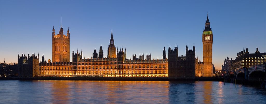 The illuminated Gothic Revival Palace of Westminster extends along the river at dusk, with several towers and the clock tower reflected in dark water.