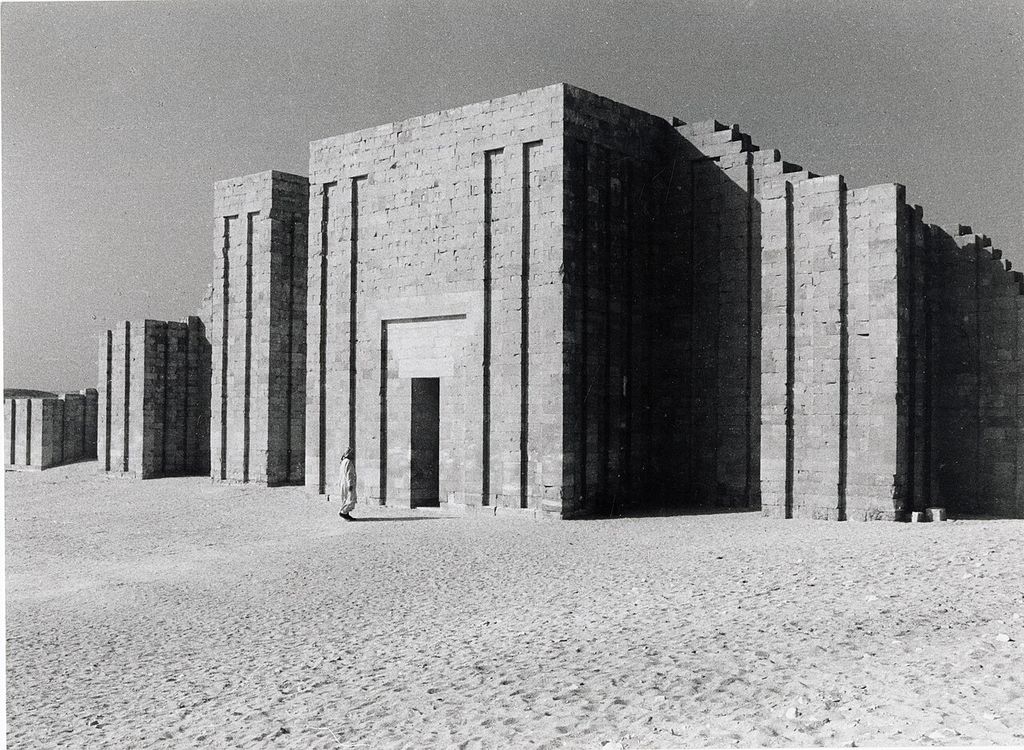 A grainy black-and-white photograph shows a very high pale stone enclosure wall with deep repeated rectangular niches and a small person at its doorway.