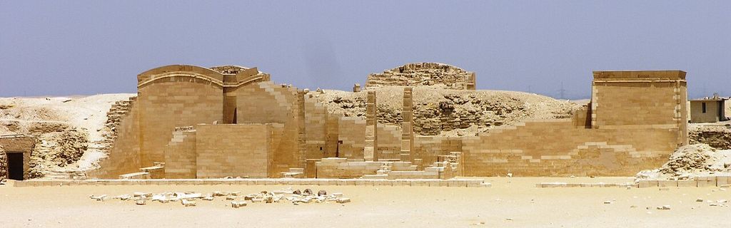 A wide desert view shows low pale stone shrine façades and court remains with the eroded stepped pyramid rising behind them.