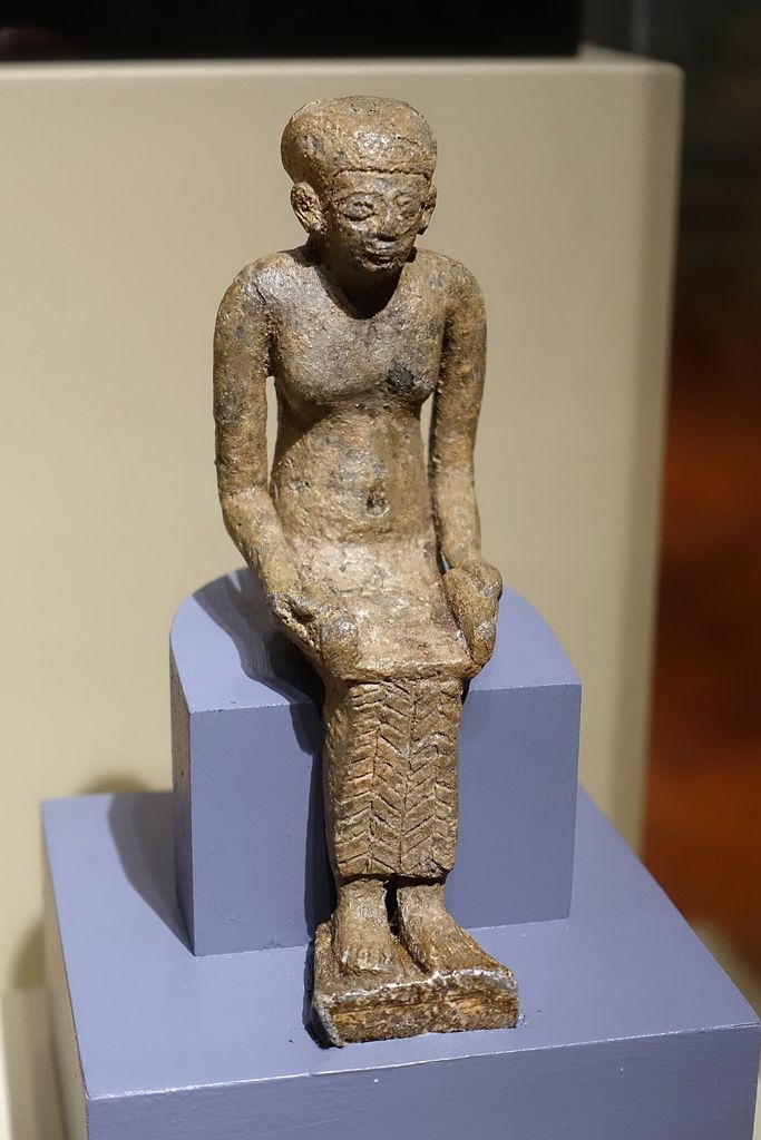A small worn bronze figure sits upright on a museum mount, wearing a close cap and long patterned garment while holding a papyrus across the lap.