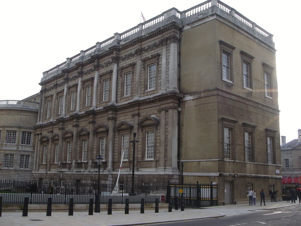An oblique street view shows a long two-storey classical façade with repeated windows, pilasters, columns and a roof balustrade beside a plain end wall.