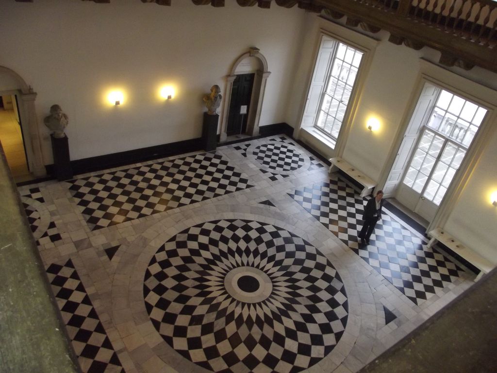A high view into a square, two-storey hall shows a black-and-white geometric floor with a large circular centre, tall windows, doors and a surrounding gallery.