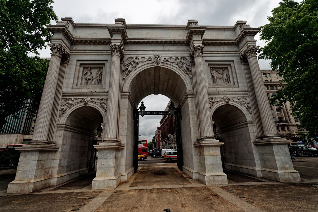 A freestanding white marble triumphal arch has one tall central opening, two smaller side arches, fluted columns and sculpted relief panels, with traffic visible through it.