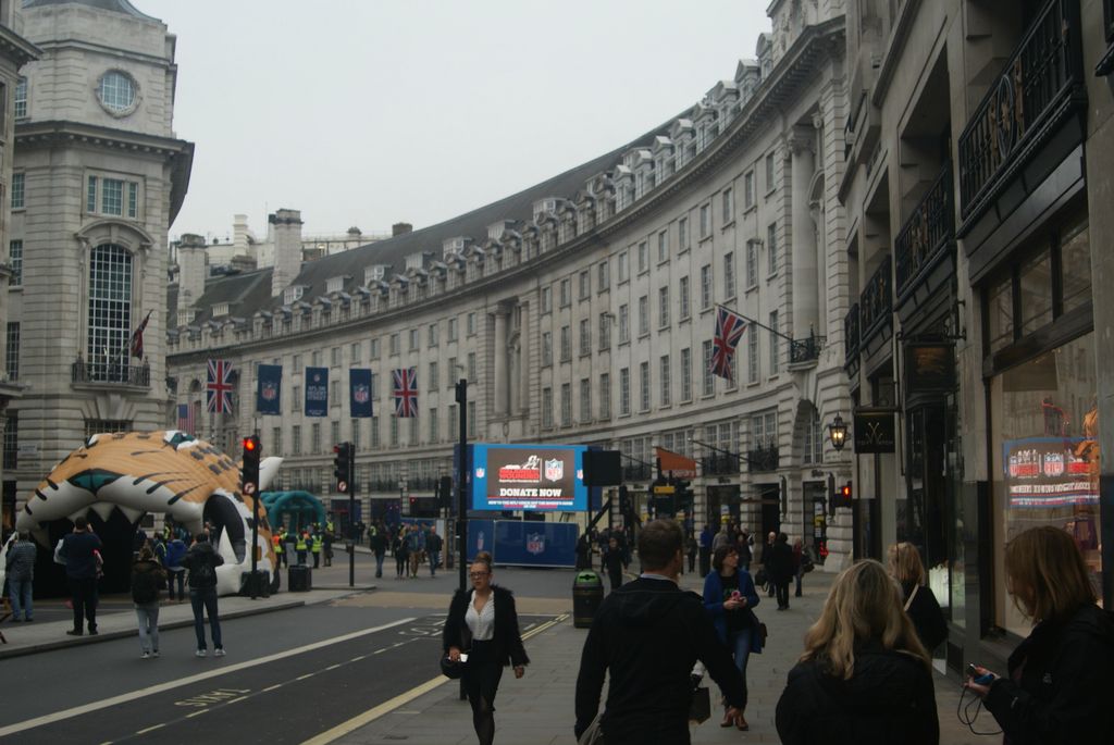 Later pale-stone commercial terraces sweep around a broad curved London street filled with pedestrians, flags, signs, a digital display and a temporary inflatable object.
