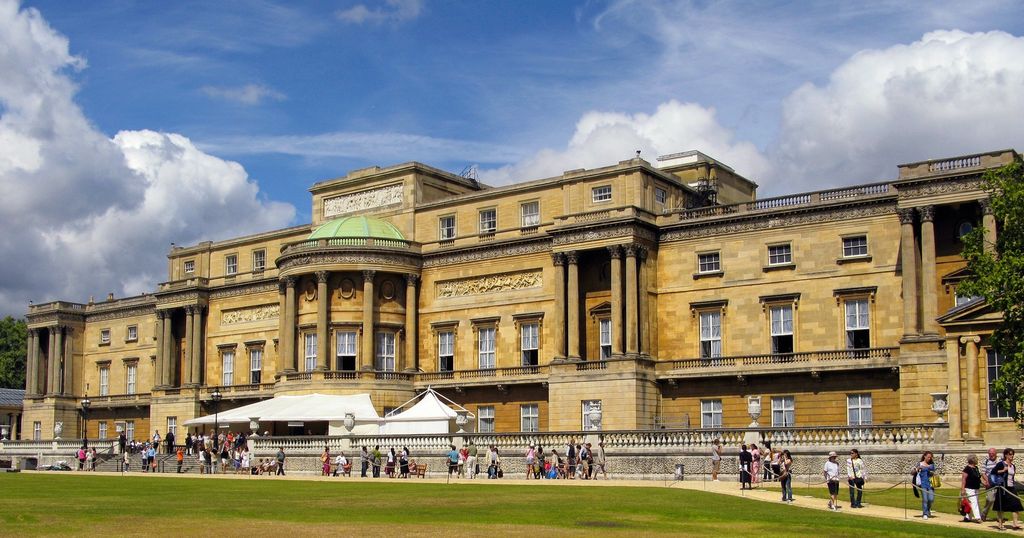 A long honey-coloured palace façade faces a broad lawn, with giant pilasters, projecting columned wings, sculpted panels, a central rounded bay, temporary tents and many visitors.