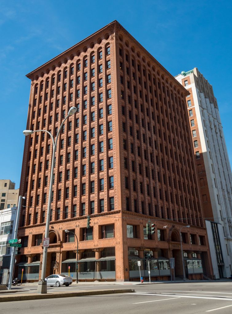 A reddish terracotta office tower rises from a richly ornamented base in closely spaced vertical piers and dark windows, ending in a row of rounded upper arches beneath a projecting cornice.