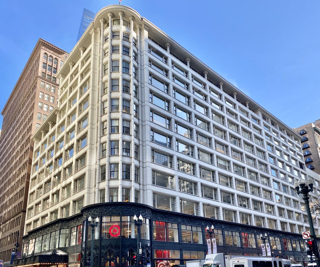 A large white-grid department store wraps a rounded Chicago corner above two dark ornamented cast-iron street levels, with big retail windows, Target signs, vehicles and a reconstructed roof loggia.