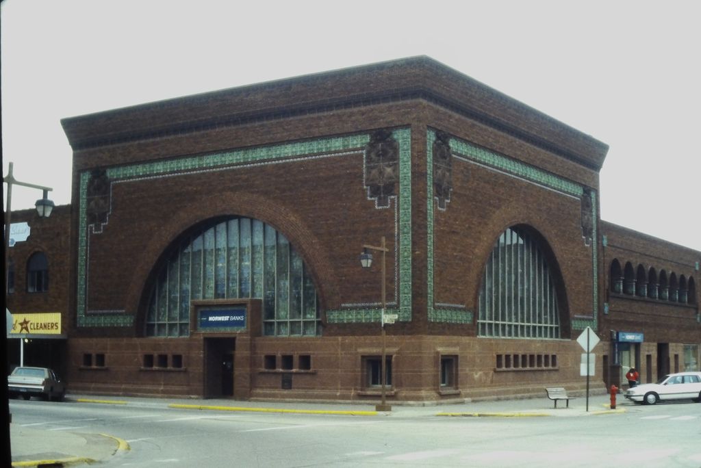 A low square red-brick bank has two enormous arched windows and turquoise-green ornamental bands around its upper walls and corners, with 1990s cars and street signs nearby.