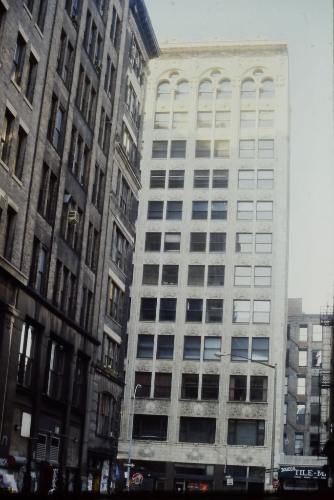 A narrow pale twelve-storey façade rises between dark neighbouring buildings, with vertical piers, broad window bays and an ornate top row of paired arches.