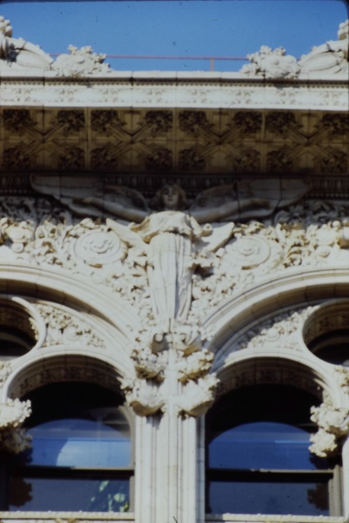 A pale winged terracotta figure spans a narrow façade pier between paired arches, surrounded by dense leaves, scrolls and geometric relief below a projecting cornice.