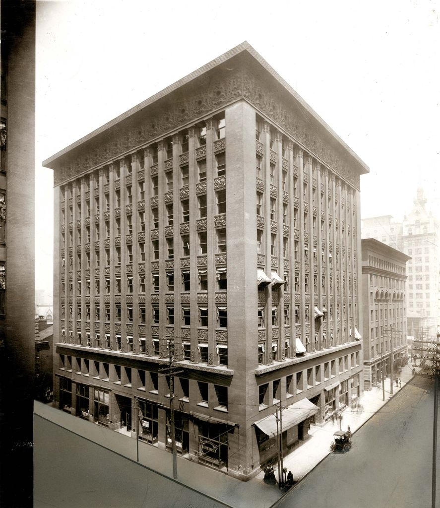 A 1907 black-and-white view shows a tall corner office building with a broad two-storey base, many narrow vertical window bays and a deep ornamental cornice above street awnings and a horse-drawn vehicle.