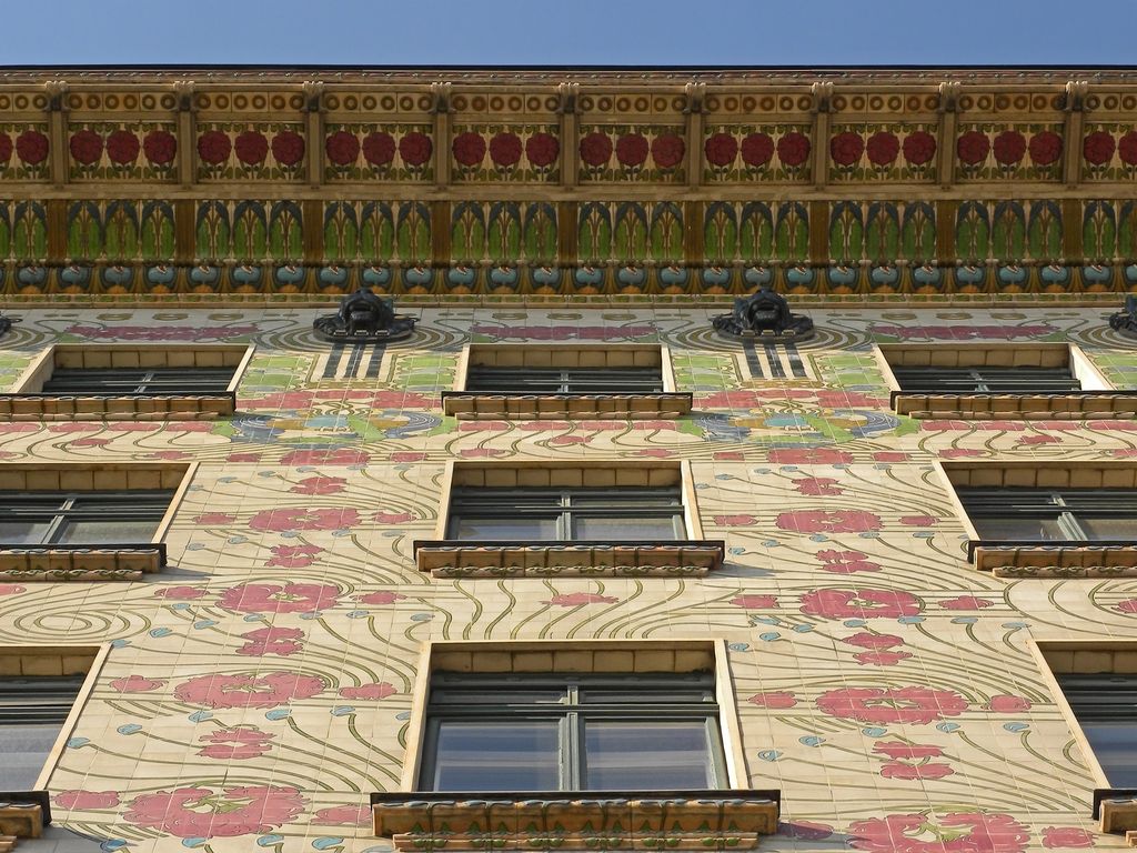 Cream ceramic tiles patterned with large pink flowers and green stems cover an apartment façade beneath a richly coloured cornice, with dark windows and shallow metal balconies.