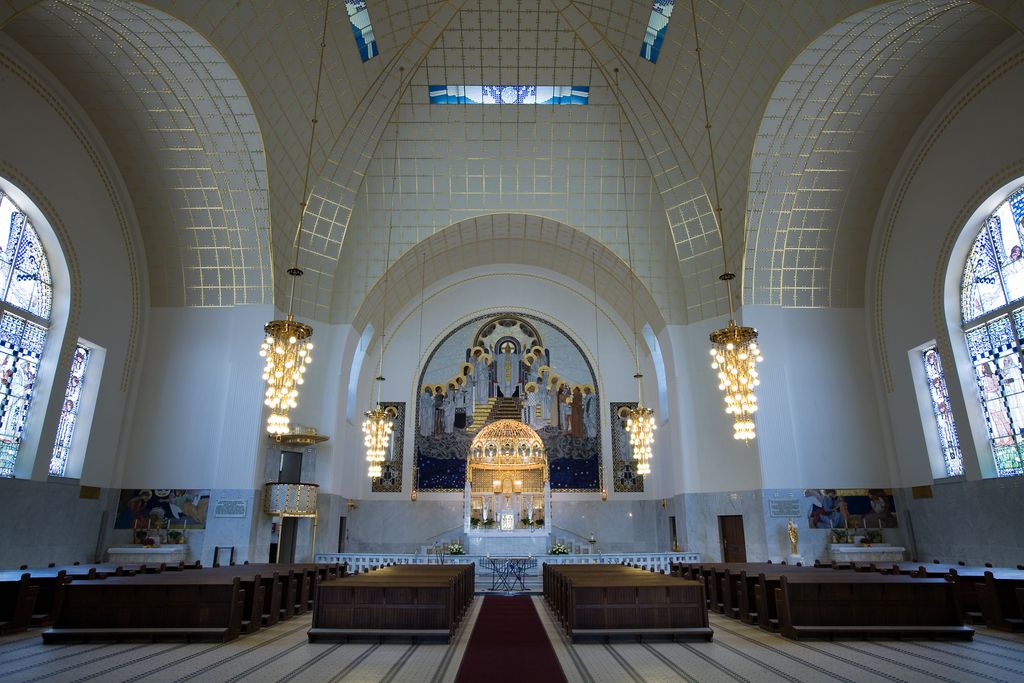 A bright white church interior has a shallow vaulted ceiling, stained-glass windows, short dark pew sections and a glowing gold altar beneath a large mosaic.
