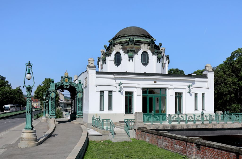 A white domed pavilion beside a railway and road has green doors, railings, ornate lamps and an elaborate separate green entrance gate.