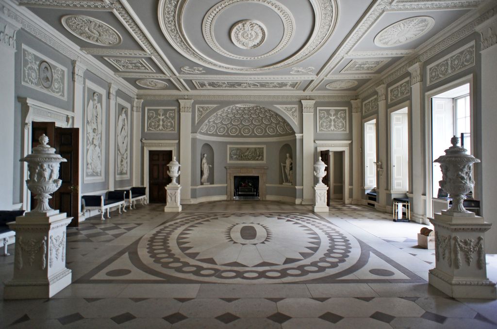 A broad grey-and-white classical hall aligns a circular patterned stone floor with an apsed fireplace wall, statues, urns, doorways and a richly coffered ceiling.