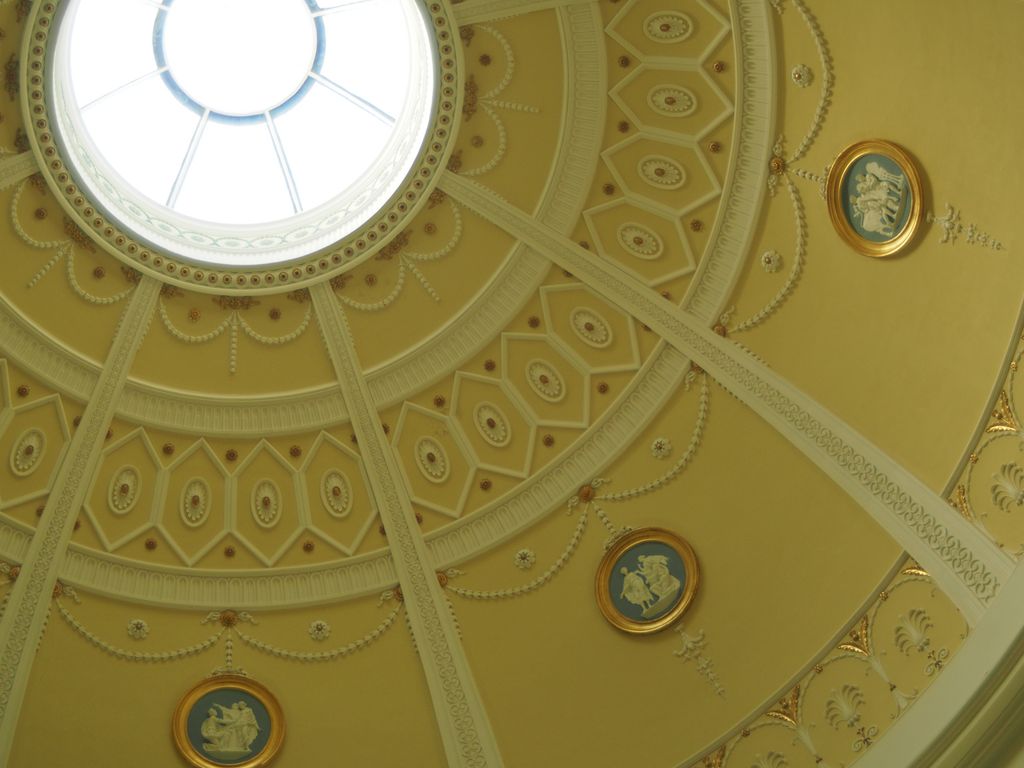 An oblique upward view of a pale yellow dome shows a bright circular skylight, radial white ribs, geometric coffers, swags, rosettes and framed classical medallions.