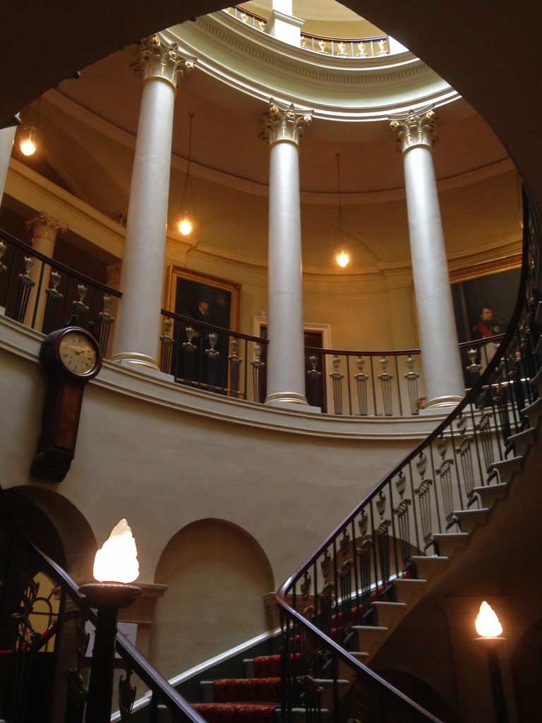 Curving stair flights and dark iron balustrades rise around tall pale Corinthian columns towards a circular gallery and bright skylight in an oval hall.