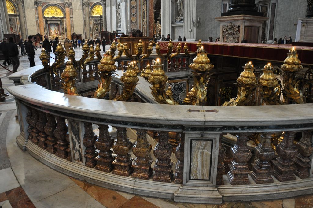 A curved marble balustrade and gilded lamps surround a recessed opening before the altar, with visitors and the base of the baldachin behind.