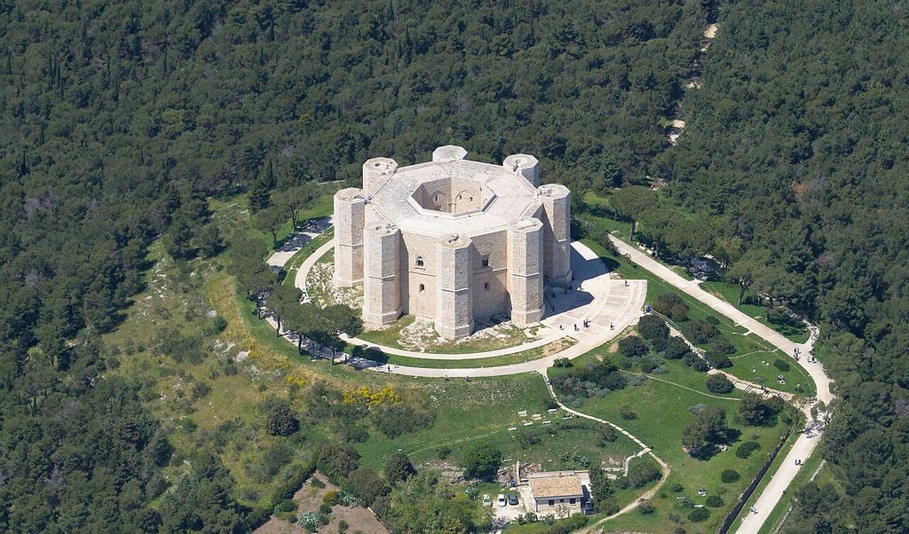 An octagonal stone building with corner towers surrounds an open court on a wooded hill.