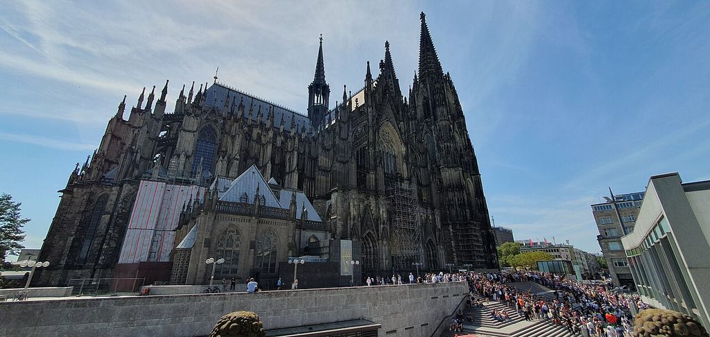 An oblique view shows a cathedral’s tall spires, transept facade, buttresses and scaffolding above a busy urban terrace.