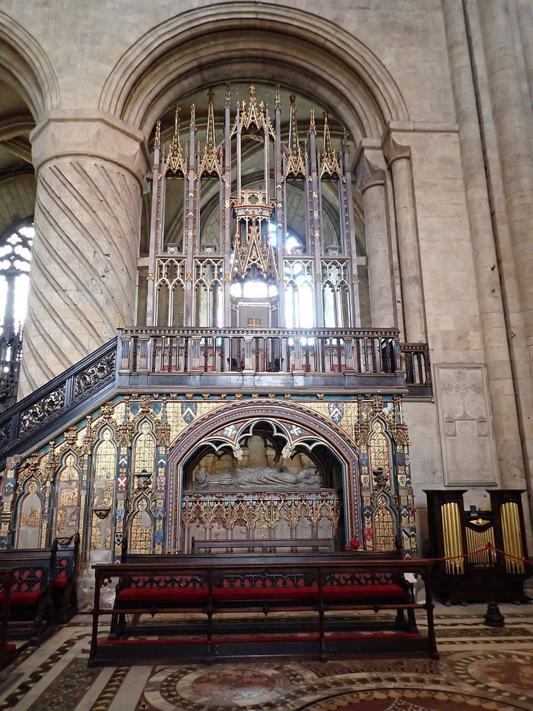 A tall decorated canopy rises over an elevated seat and balustrade above a stone tomb.