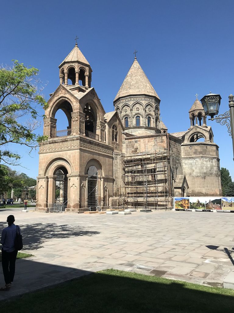 A conical dome on a windowed drum rises behind an arched tower and stone walls with scaffolding.