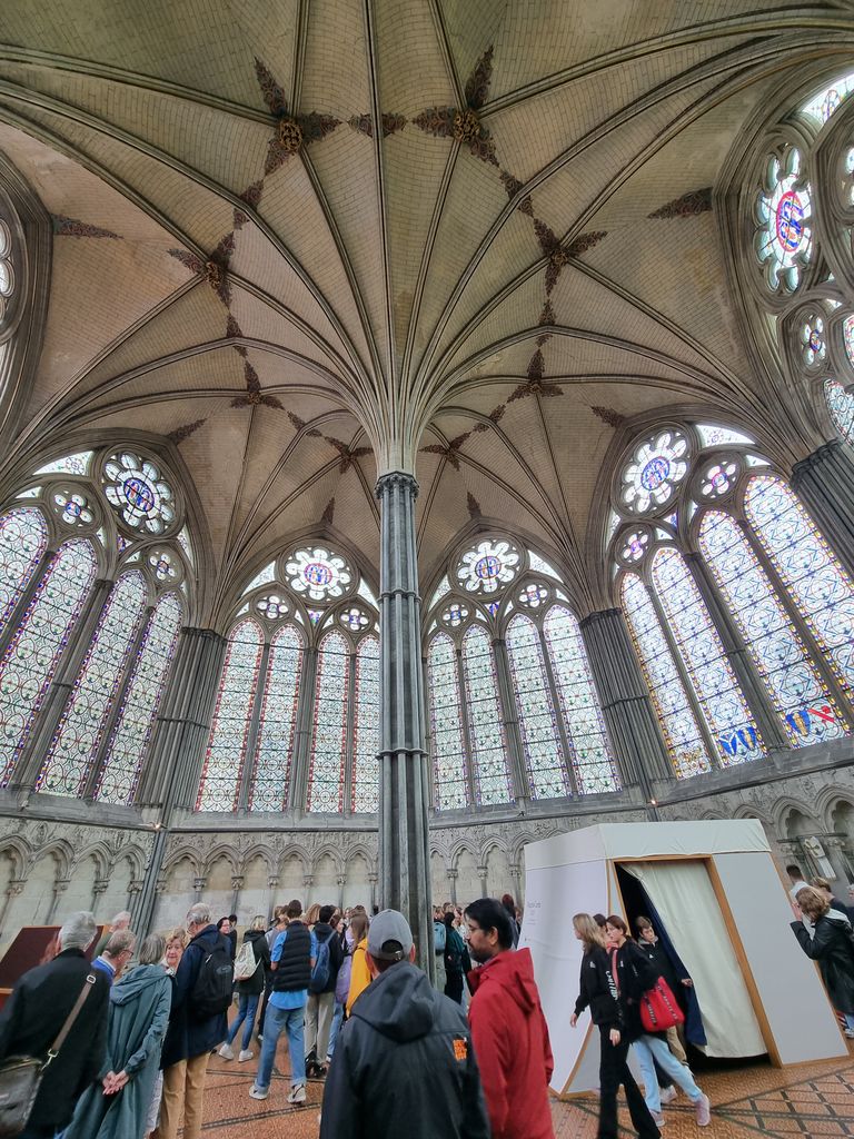 Vault ribs spread from a slender central support above a room lined with large traceried windows, visitors and an exhibition booth.