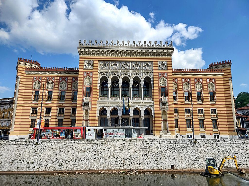 A striped red and ochre civic façade with pointed arches and carved geometric panels stands behind a tram beside a river wall.