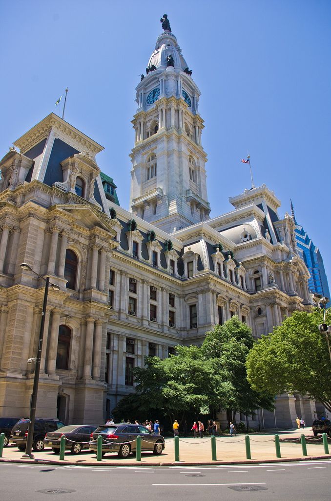 An immense pale masonry city hall rises in layered pavilions beneath a central clock tower, with cars, trees and pedestrians at street level.