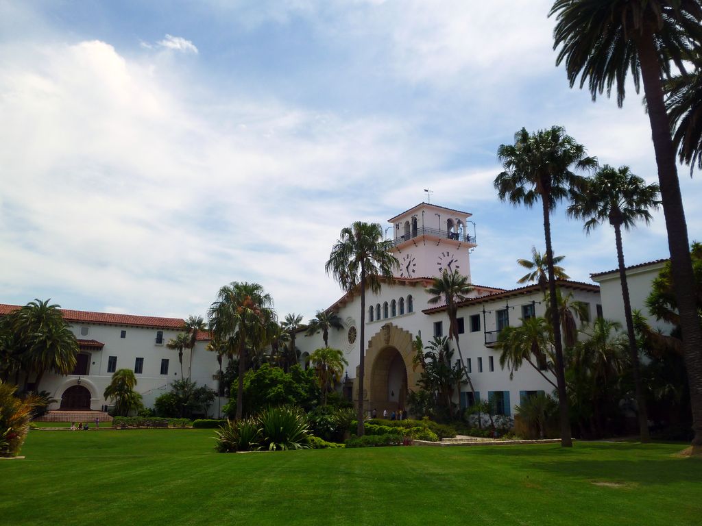 White stucco courthouse wings, arched openings, tiled roofs and a clock tower stand beyond palms and a broad green garden.