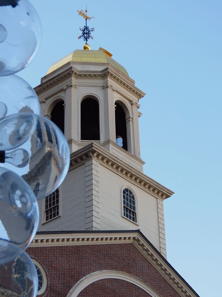 A brick gable rises to a white clock stage, open arched cupola and gilded grasshopper weather vane beside a cropped foreground sculpture.