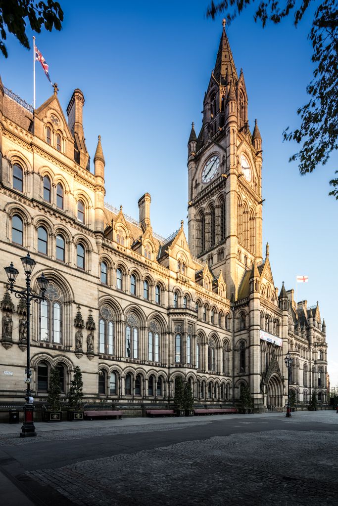 A long Gothic Revival civic façade with arcades, traceried windows, steep roofs and a tall clock tower borders a paved square.